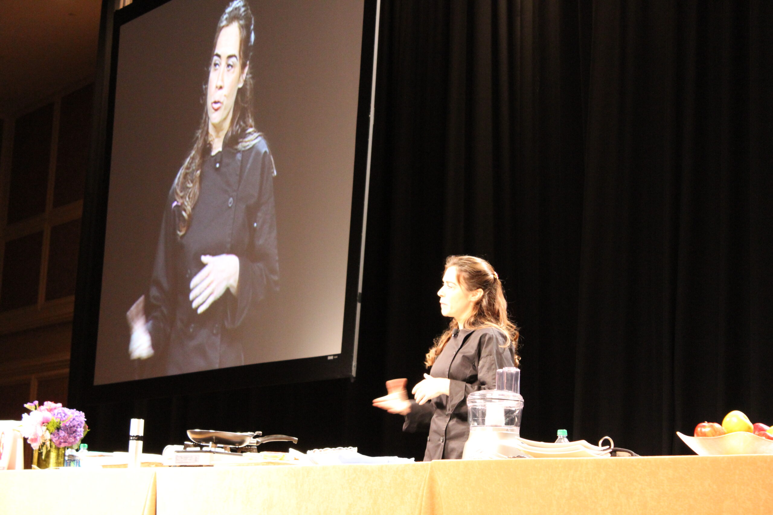 Devin Alexander on stage running a live cooking demo with her image projected behind her